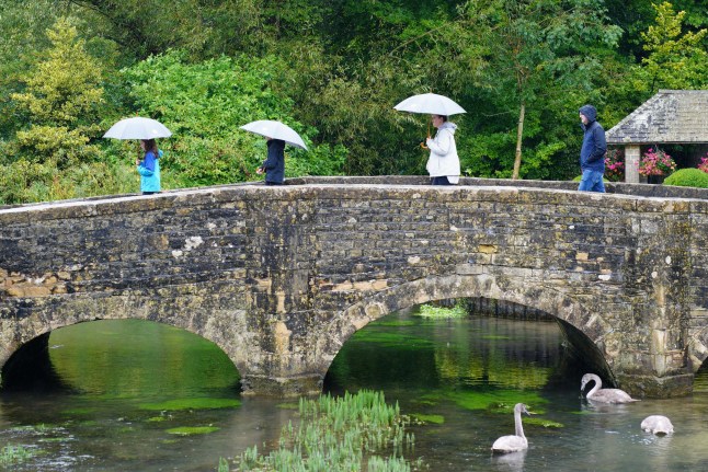 Visitors with umbrellas in the rainy weather at Bibury village in Gloucestershire. Picture date: Saturday August 24, 2024. PA Photo. Photo credit should read: Ben Birchall/PA Wire