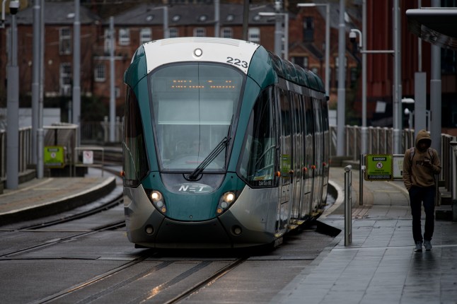 Nottingham Express Transport NET Tram approaches the Nottingham Station stop on a rainy day as a hooded young man walks alongside looking at his phone.