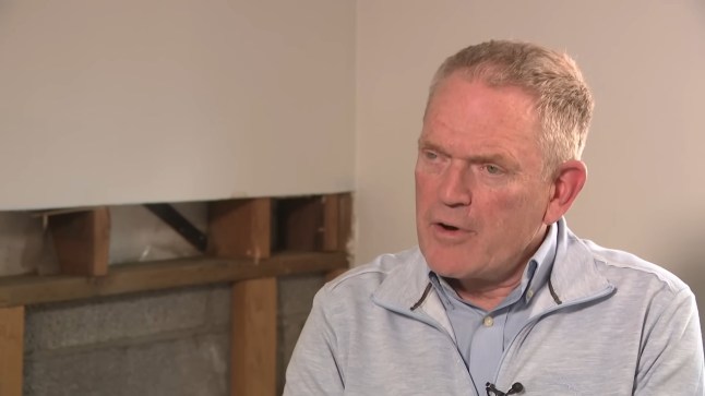 A man wearing a grey shirt speaking ahead of an exposed patch of wall.