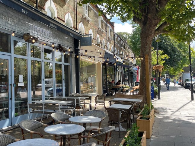 A promenade of shops in a leafy London street is seen on a warm Monday afternoon. An exterior of several shops are seen with their tables and chairs outside