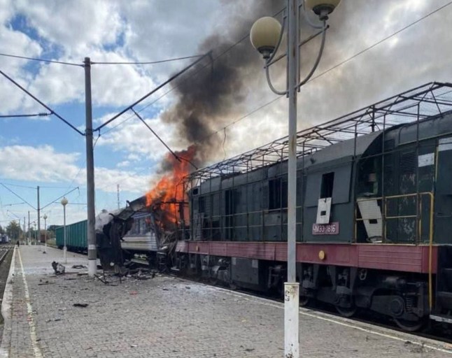 A train carriage engulfed in orange flames and black smoke billows from the train after being struck by a Russian drone