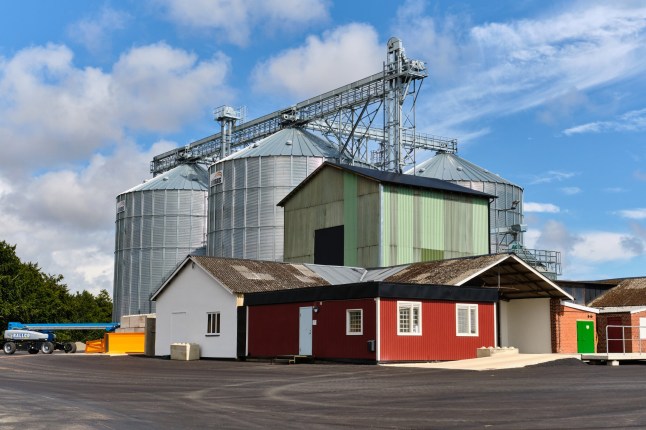 July 23, 2025 ?????? Ystad-Sweden: Recently constructed grain silos rise behind older farm buildings at an agricultural storage and feed facility under a partly cloudy summer sky.