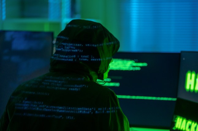 Rear view of a hooded hacker organizing a massive data breach attack on corporate servers in a dark, secret location. A computer hacker is coding in a secret location, using multiple screens to bypass firewalls and security systems.