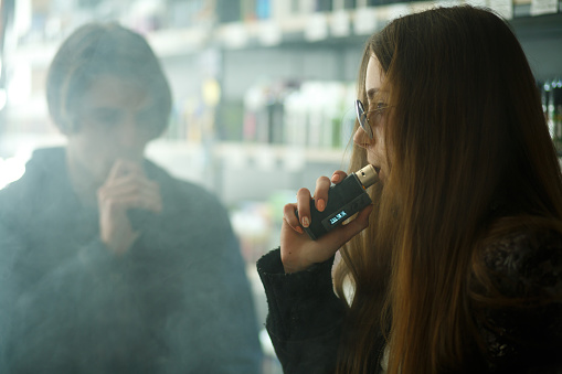Vape teenagers. Young cute girl in sunglasses and young handsome guy smoke an electronic cigarettes in the vape bar. Bad habit. Vaping activity.