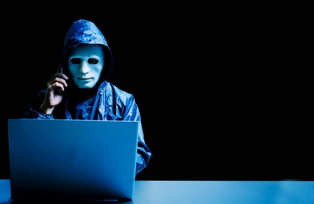 Anonymous computer hacker in white mask and hoodie. Obscured dark face using laptop computer for cyber attack and calling on cellphone, Data thief, internet attack, darknet and cyber security concept.; Shutterstock ID 1438588631; purchase_order: -; job: -; client: -; other: -