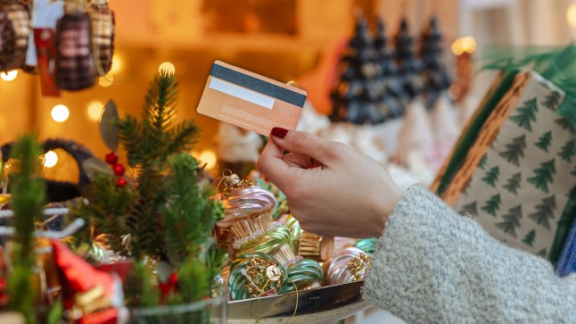 A joyful winter market scene with a smiling woman in a red beanie and blue scarf, selecting a gift card among holiday gifts and twinkling lights.