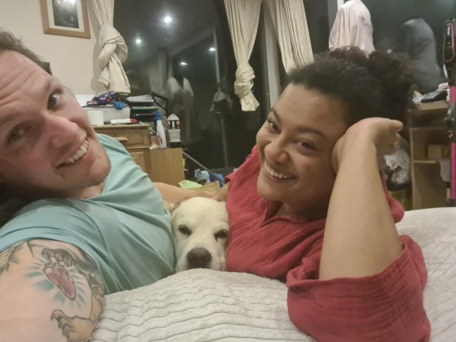 A couple lying on their bed with their dog sandwiched between them while they smile for the camera